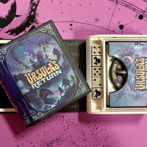 May include: A white box with a black and blue book lid featuring the text "Ursula's Return" and a fantasy illustration. The box also contains a small, white, six-sided die and a round, black and white dial with the text "Lorcana" on it.