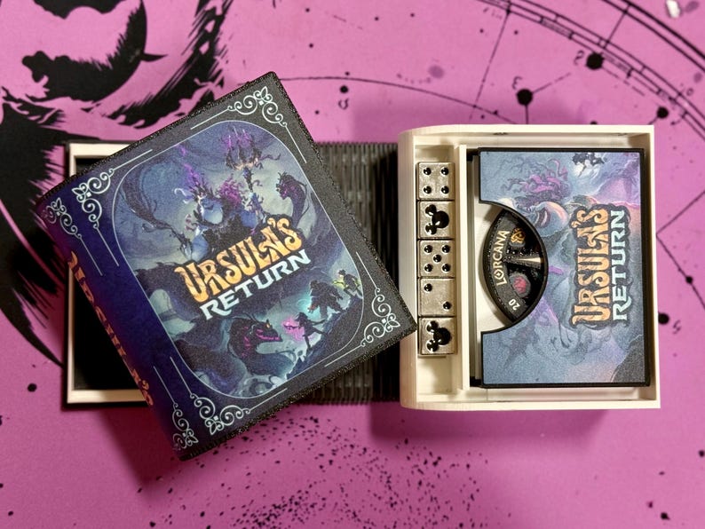 May include: A white box with a black and blue book lid featuring the text "Ursula's Return" and a fantasy illustration. The box also contains a small, white, six-sided die and a round, black and white dial with the text "Lorcana" on it.