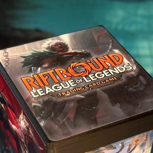 May include: A square game box featuring the title RIFTBOUND LEAGUE OF LEGENDS TRADING CARD GAME in orange and white text. The lid displays a character illustration with white hair and dark armor, while the side shows red and black artwork.