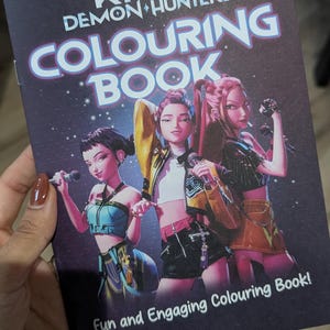 Op de afbeelding: Een KPOP Demon Hunters kleurboek met een donkerpaarse omslag. De omslag toont drie geanimeerde vrouwelijke figuren die microfoons vasthouden. De tekst "COLOURING BOOK" is prominent aanwezig. De zin "Fun and Engaging Colouring Book!" is ook zichtbaar.