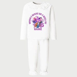 May include: White pajama set with a long-sleeved top and pants. The top features a round collar with lace trim and a graphic with the text "WHEN I WAKE UP I WILL BE 6 NAME". The graphic includes cartoon characters in purple, pink, and blue.