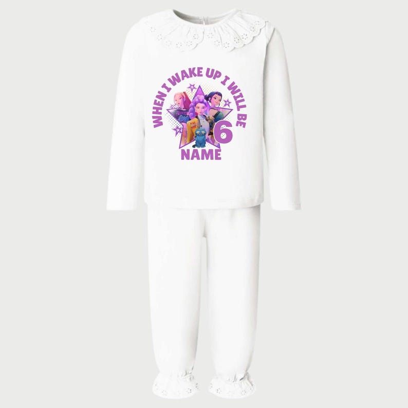 May include: White pajama set with a long-sleeved top and pants. The top features a round collar with lace trim and a graphic with the text "WHEN I WAKE UP I WILL BE 6 NAME". The graphic includes cartoon characters in purple, pink, and blue.