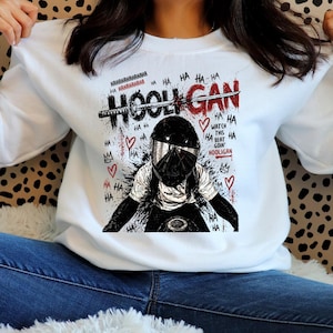 May include: A white crewneck sweatshirt featuring a graphic print of a person wearing a black motorcycle helmet. The design includes the text HOOLIGAN, HA HA, and WATCH THIS BEAT GOIN HOOLIGAN, along with red hearts, crowns, and a sword illustration.