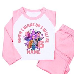 May include: Pink and white pajama set with a long-sleeved top and matching pants. The top features a graphic of three cartoon characters, a blue cat, and the text "WHEN I WAKE UP I WILL BE 6 NAME".