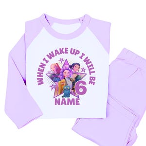 May include: A light purple and white pajama set. The long-sleeved top features a graphic of three animated girls, a blue cat, and the text "WHEN I WAKE UP I WILL BE 6 NAME". The matching pants are light purple.