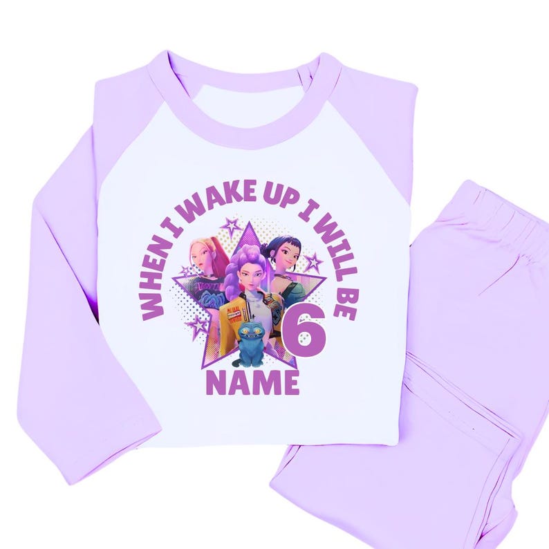 May include: A light purple and white pajama set. The long-sleeved top features a graphic of three animated girls, a blue cat, and the text "WHEN I WAKE UP I WILL BE 6 NAME". The matching pants are light purple.