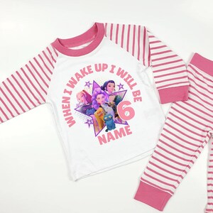 May include: White and pink striped pajama set. The long-sleeved top features a graphic of animated characters and the text "WHEN I WAKE UP I WILL BE 6 NAME". The sleeves and pants are striped pink and white.