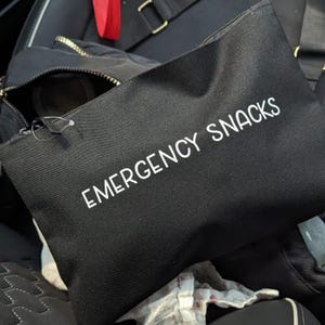 May include: Black zippered pouch with the words "EMERGENCY SNACKS" printed in white. The pouch is made of a textured fabric and is slightly angled. The background includes a partially visible black bag and other items.