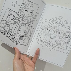 Op de afbeelding: Een open kleurboek met zwart-wit illustraties van stripfiguren. Het boek toont tekeningen met de tekst "KPOP MON HUNTERS" en "Six Boys POPS". Een hand, met een lichtpaars horloge, houdt de pagina's open.