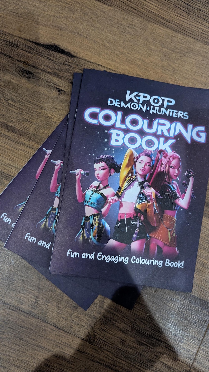 Op de afbeelding: Verschillende exemplaren van een kleurboek getiteld "KPOP Demon Hunters Colouring Book" liggen op een houten oppervlak. De cover toont drie geanimeerde figuren met microfoons. De tekst "Fun and Engaging Colouring Book!" is ook zichtbaar.