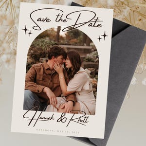 Puede incluir: Tarjeta de Save the Date con una foto de una pareja en un entorno boscoso. La tarjeta tiene un fondo blanco con texto negro que dice "Save the Date" y "Hannah & Rhett". La fecha está escrita debajo de los nombres como "SÁBADO, 18 DE MAYO DE 2023".