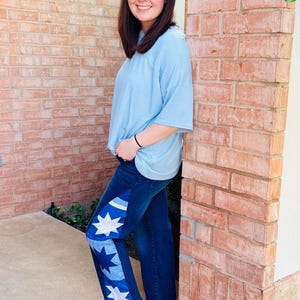 May include: A light blue t-shirt and dark wash denim jeans with star-shaped patches. The jeans have a cropped, wide-leg fit. The person is wearing brown sandals with gold buckles. The background is a red brick wall.