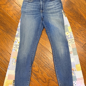 May include: High-waisted blue denim jeans featuring a straight-leg cut and raw, frayed hems. The outer side seams are opened and inset with a wide, vertical panel of multi-colored patchwork fabric, showcasing various floral, gingham, and lace patterns.