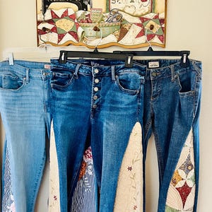 May include: Three pairs of blue denim jeans hang on a rack. Each pair features unique decorative panels, including floral patterns, quilted designs, and embroidered details. A sign above reads "Sew Cute."