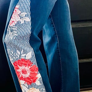 May include: Blue denim jeans with a floral and striped side panel. The panel features red and peach-colored flowers with green leaves on a white and blue striped background. The jeans have a classic five-pocket design.