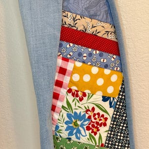 May include: A light blue denim jacket with a patchwork design on the right side. The patchwork is made up of various colorful fabrics with floral, geometric, and polka dot patterns.
