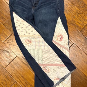 May include: Dark blue denim jeans with patchwork fabric panels down the sides. The patchwork features floral and geometric patterns in shades of pink, cream, and green. The jeans have a classic five-pocket design.