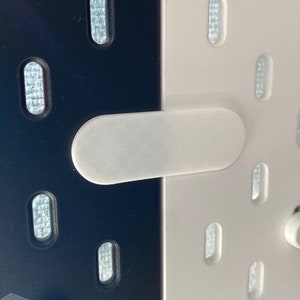 Könnte beinhalten: Eine weiße Kunststoffplatte mit einer Reihe ovaler Löcher. Eine weiße rechteckige Kunststoffplatte ist an der Platte befestigt und bedeckt einige der Löcher.