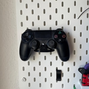 Könnte beinhalten: Ein schwarzer PlayStation 4-Controller ist an einer weißen Lochwand befestigt. Der Controller hat farbige Aktionstasten und ein Touchpad. Ein schwarzer Halter ist an der Lochwand befestigt, um den Controller zu halten. Weiteres Zubehör ist ebenfalls montiert.