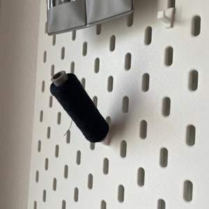 Könnte beinhalten: Eine schwarze Garnrolle hängt an einer weißen Lochwand mit gleichmäßig verteilten Löchern.