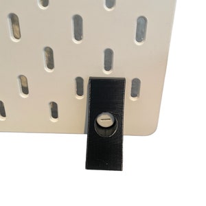 Könnte beinhalten: Ein schwarzer Kunststoff-Pegboard-Haken mit einem Loch in der Mitte. Der Haken ist an einer weißen Lochwand befestigt.
