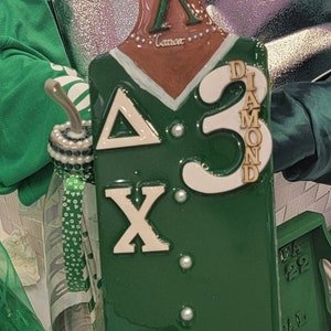 Op de afbeelding: Een groen en wit houten peddel met de letters "Delta Sigma Theta" en het nummer "3" erop. De peddel heeft ook de woorden "Diamond" en "7 Jewels" erop. De peddel is versierd met gouden glitter en witte parels.