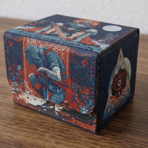 May include: A rectangular deck box featuring a textured surface with a dark blue and red illustration. The front panel depicts a kneeling figure, while the side panel displays a triangular emblem with a central eye motif against a dark background.