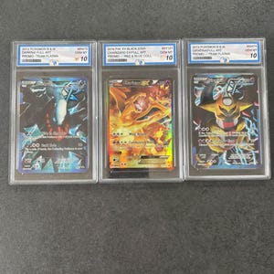 May include: Three graded Pokémon trading cards in protective cases. The cards feature Charizard, Darkrai, and Giratina. Each card is graded GEM MT 10. Text on the cards includes "Team Plasma" and "Full Art". The Charizard card is from the XY Black Star series.