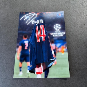 May include: A navy blue football shirt with the number 14 in red, and the name "ODOU" on the back. The shirt is hanging on a pole in front of a blurred football pitch. The Champions League logo is visible.