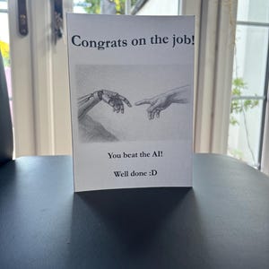 May include: A white greeting card featuring the text "Congrats on the job!" above a black and white illustration of a robotic hand and a human hand. The card also reads "You beat the AI!" and "Well done :D".