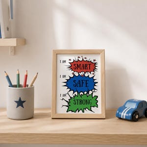 Könnte beinhalten: Ein gerahmter Druck mit drei Sprechblasen im Comic-Stil in Rot, Blau und Grün. Der Text lautet I AM SMART, I AM SAFE und I AM STRONG. Der Druck befindet sich in einem hellen Holzrahmen, begleitet von einem grauen Stiftehalter und einem blauen Spielzeugauto.
