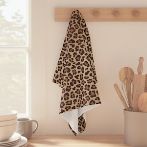 May include: A rectangular kitchen towel featuring a repeating leopard print pattern in shades of tan, brown, and black. The towel is draped over a wooden wall-mounted rack, displaying a soft, textured fabric surface with a white underside visible.