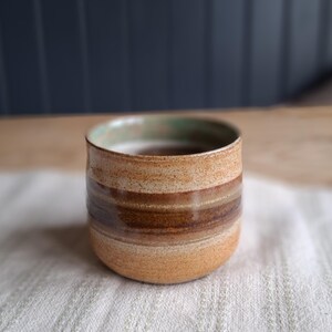 May include: A handcrafted ceramic tumbler with a rounded base and a cylindrical body. The cup features horizontal bands of tan, brown, and cream colours. The rim has a subtle green hue. The tumbler is likely used for drinking beverages.