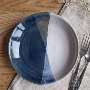 May include: A handcrafted ceramic plate with a two-tone design. Half of the plate is a deep blue, the other half is creamy white. The plate is on a wooden surface, with silverware and a linen napkin.