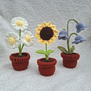 Könnte beinhalten: Drei gehäkelte Blumenarrangements in Terrakotta-Töpfen. Die Blumen umfassen Gänseblümchen mit weißen Blütenblättern und gelben Zentren, eine Sonnenblume mit gelben Blütenblättern und einem braunen Zentrum sowie Glockenblumen.