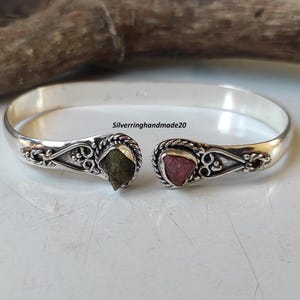 May include: A silver cuff bracelet with decorative ends. Each end features a natural gemstone, one green and one pink, set in a detailed silver setting. The bracelet is open, allowing for easy wear.