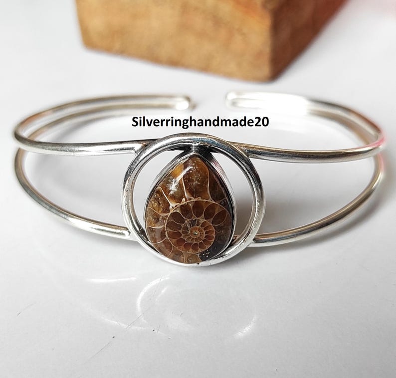 May include: Silver cuff bracelet featuring a teardrop-shaped fossil set in a circular frame. The fossil displays a detailed spiral pattern in shades of brown. The bracelet has a double-band design and the text "Silverringhandmade20".