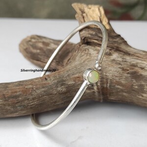 Puede incluir: Un brazalete de plata con una sola piedra redonda de ópalo. El ópalo es de color verde pálido con un ligero brillo iridiscente.