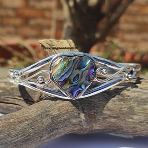 Puede incluir: Un brazalete de plata con un corazón de nácar de abulón. La concha tiene colores iridiscentes de azul, verde y morado.