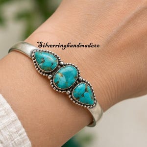 May include: A silver cuff bracelet featuring three turquoise stones with brown matrix patterns. The centre stone is oval, flanked by two pear-shaped stones, all set in beaded silver bezels. The text Silverringhandmade20 is visible above the bracelet.