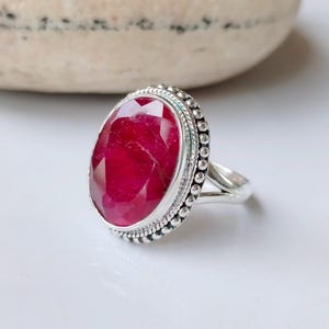 May include: A silver ring with a large, oval-shaped ruby gemstone. The ring has a beaded border around the stone.