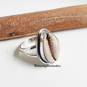 Könnte beinhalten: Ein Silberring mit einer natürlichen Kaurimuschel, die in einer polierten Metallfassung sitzt. Die Ringschiene ist in zwei dünne Silberdrähte geteilt. Der Text Silverringhandmade20 ist unten im Bild unter dem Ring schwarz aufgedruckt.