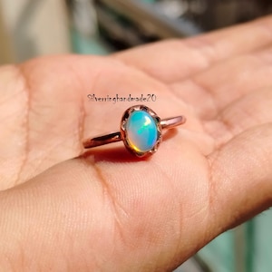 May include: A copper ring with a white opal gemstone set in a delicate, scalloped bezel. The ring is shown on a hand.