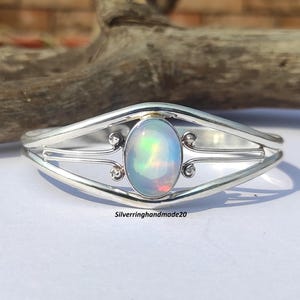 Può includere: Un braccialetto rigido in argento con un design delicato che presenta una pietra di opale ovale. L'opale ha una base bianca lattiginosa con bagliori di colore iridescente.