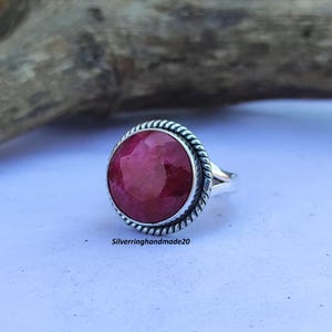 Può includere: Un anello d'argento con una grande pietra preziosa di rubino rotonda e sfaccettata incastonata in un bordo a corda.