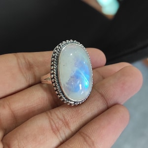 Peut inclure: Une bague en argent avec une grande pierre de lune ovale. La pierre a un reflet bleu irisé.
