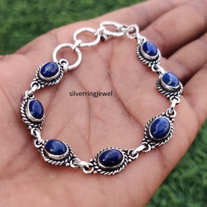 以下が含まれることがあります： チェーンリンクのデザインと楕円形のラピスラズリ宝石が付いたシルバーブレスレット。ブレスレットは留め金で留まります。