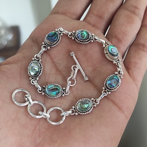 Könnte beinhalten: Ein silbernes Armband mit einem Schiebeverschluss verfügt über ovale Einlagen aus Abalone-Schale. Das Armband hat ein Gliederketten-Design.
