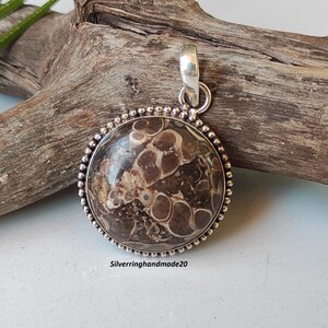 May include: A round pendant with a brown and white patterned stone set in a silver bezel with a beaded edge. The pendant is attached to a silver bail.