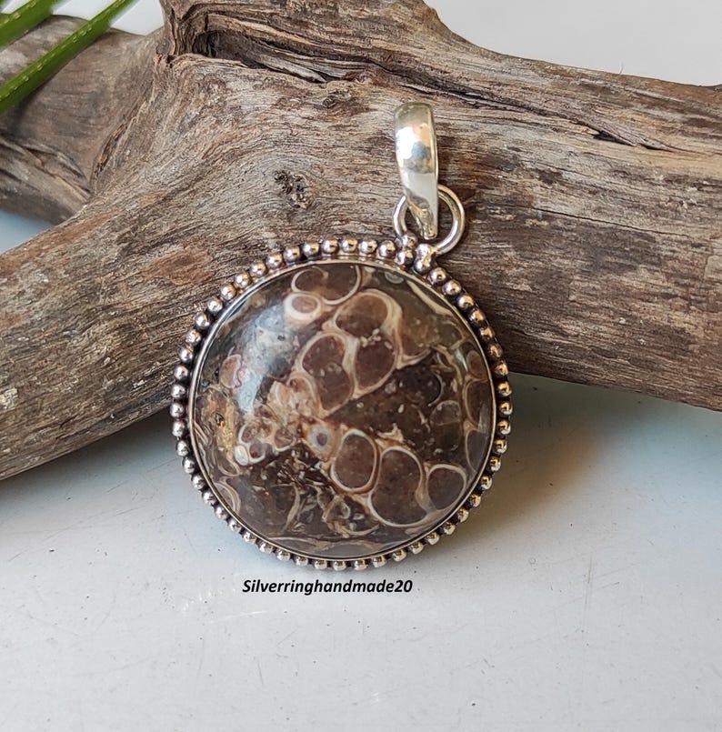 May include: A round pendant with a brown and white patterned stone set in a silver bezel with a beaded edge. The pendant is attached to a silver bail.